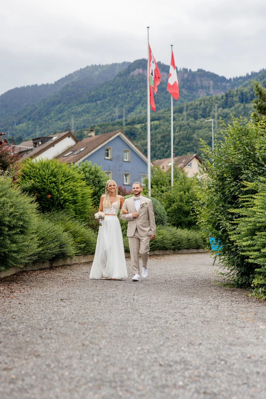 Brautpaar geht einen Kiesweg zwischen Hecken entlang, dahinter zwei Fahnenmasten und ein bewaldeter Berg