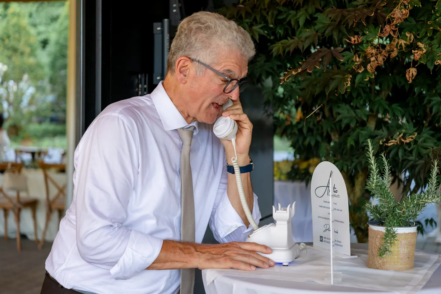 Ein älterer Herr spricht in den Hörer des weissen Telefons, daneben stehen der Aufsteller mit der Anleitung und ein Rosmarintopf