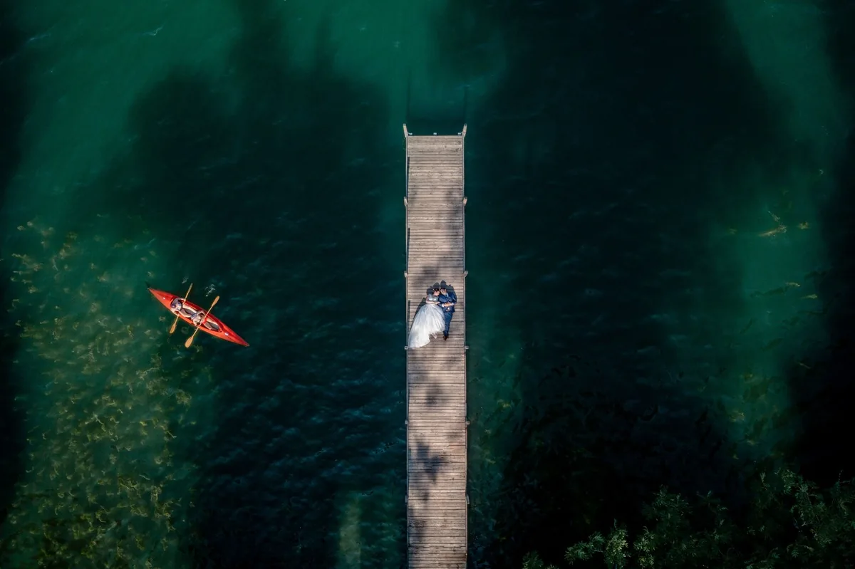 Brautpaar am Ende eines Stegs im türkisen Wasser, links fährt ein rotes Kajak vorbei