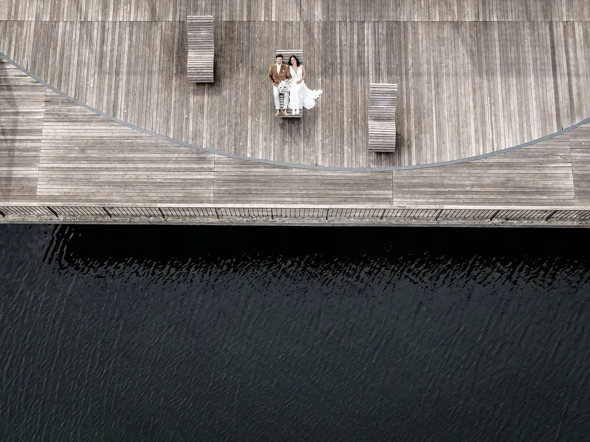 Brautpaar auf einem breiten Holzdeck am See, im Holz zeichnet sich eine grosse Bogenlinie ab, daneben zwei Bänke