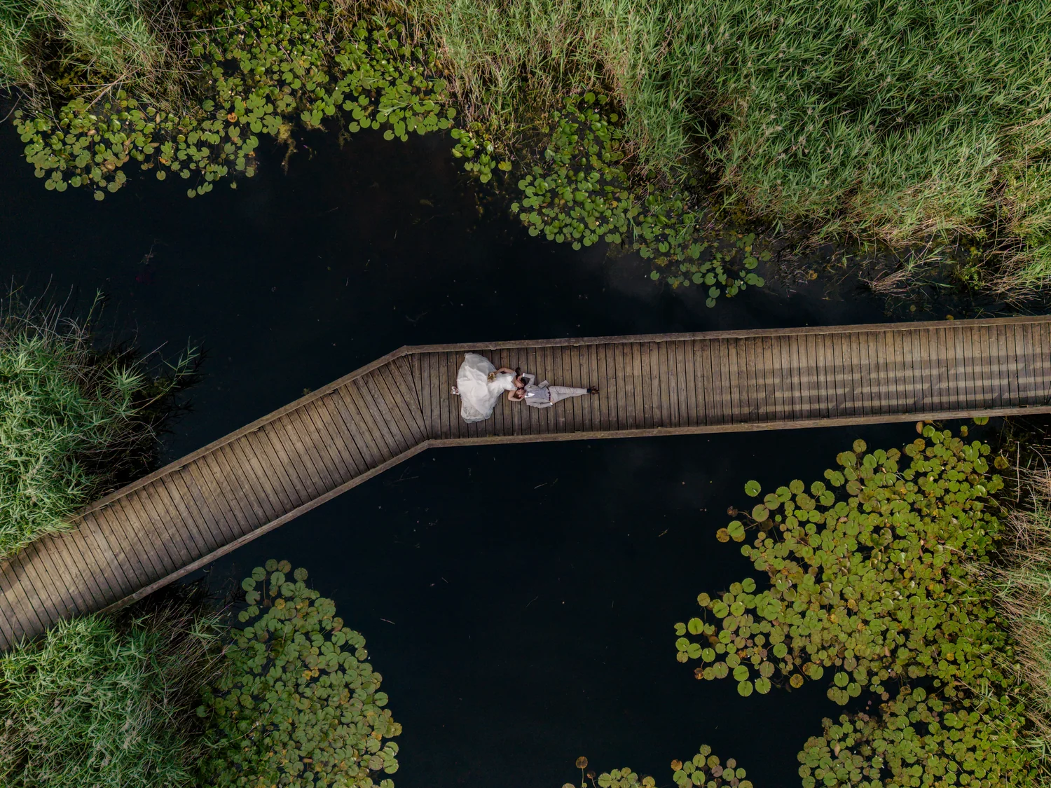 Brautpaar liegt an der Biegung eines Holzstegs über dunklem Wasser mit Seerosenblättern