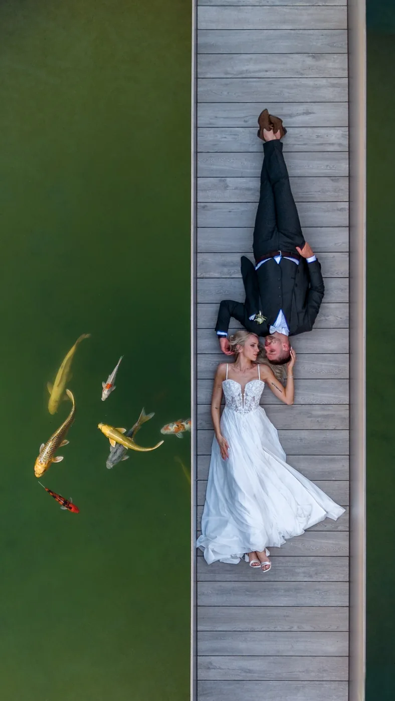 Brautpaar liegt auf dem Steg über dem Koiteich beim Heuboden in Holzhäusern, daneben ziehen die Fische durchs Wasser, senkrecht von oben