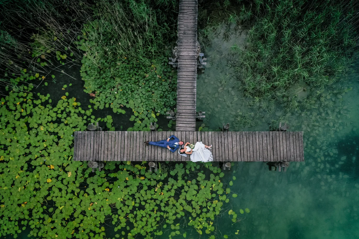 Brautpaar liegt auf einem Holzsteg in einem Weiher voller Seerosenblätter, ringsum Schilf