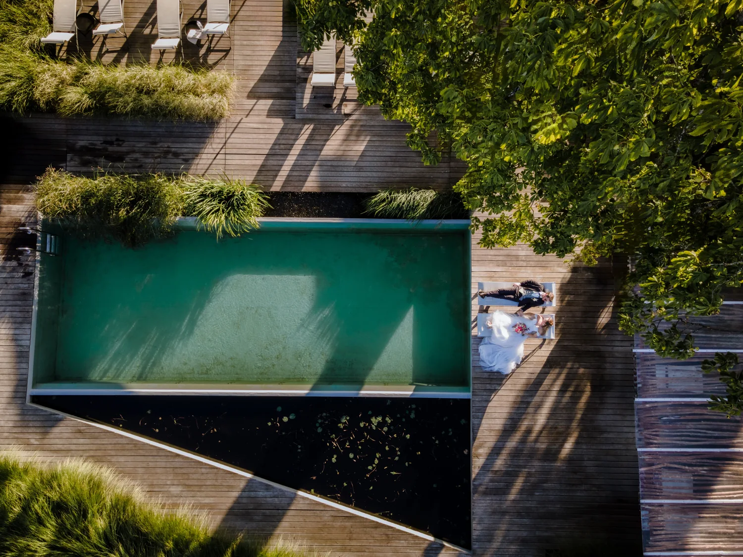 Brautpaar liegt nebeneinander auf zwei Liegen neben einem Pool, der Schatten eines Baums fällt über die Holzdielen