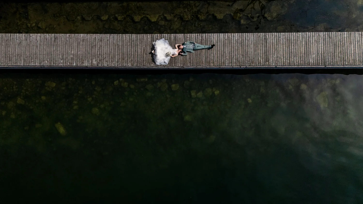 Ein Steg zieht sich quer durchs ganze Bild, in der Mitte liegt das Brautpaar, oben und unten Wasser
