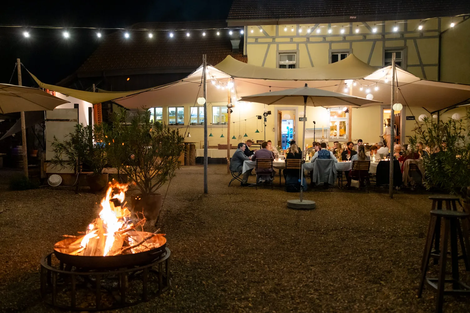 Feuerschale brennt am Abend im Hof, dahinter sitzen Gäste unter Lichterketten