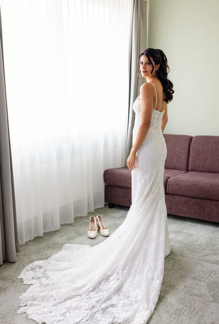 Braut im Hochzeitskleid steht am Fenster eines hellen Hotelzimmers