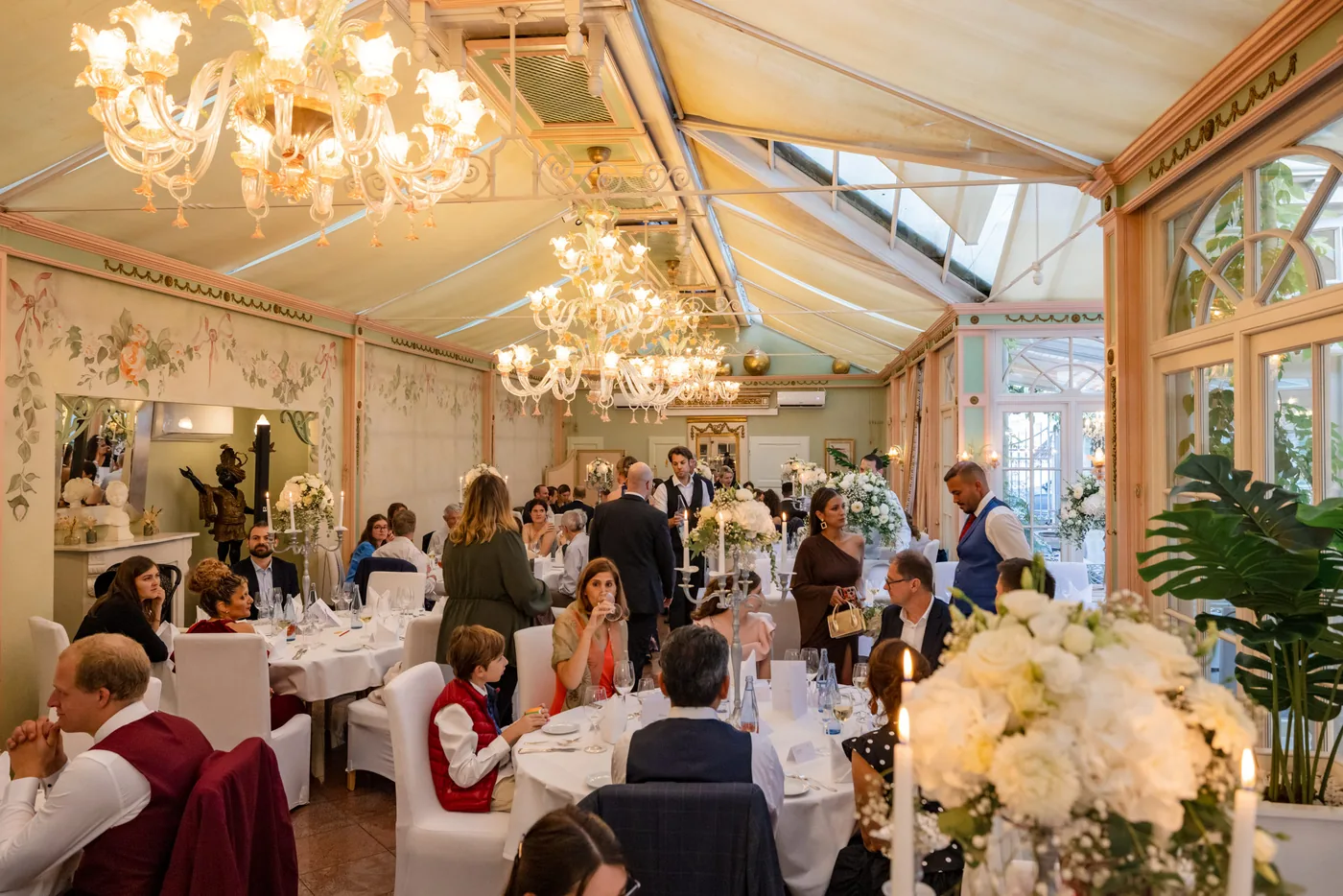 Hochzeitsgesellschaft sitzt an gedeckten Tischen im Wintergarten unter Kronleuchtern