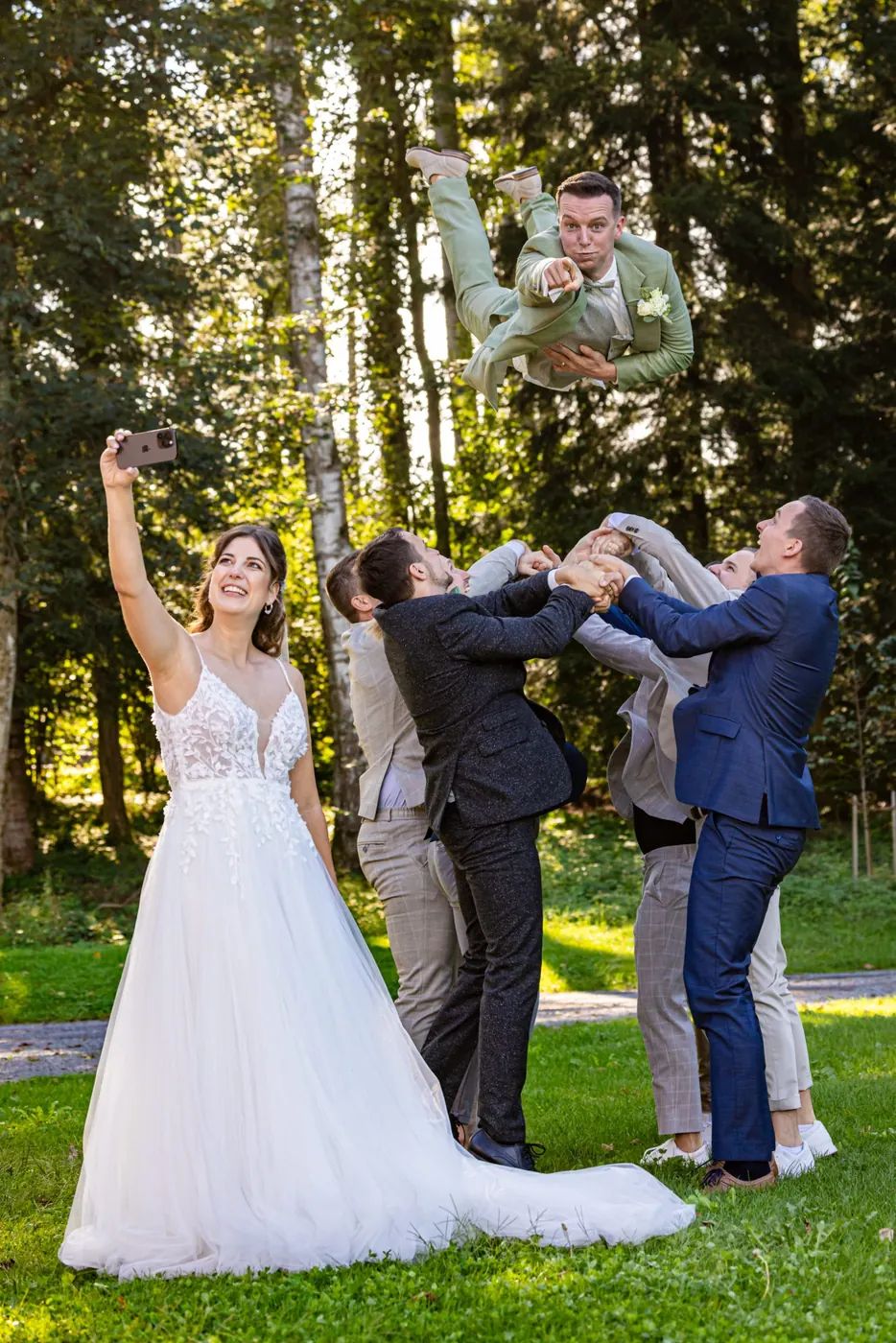 Die Braut macht ein Selfie, während hinter ihr die Freunde den Bräutigam hoch in die Luft werfen
