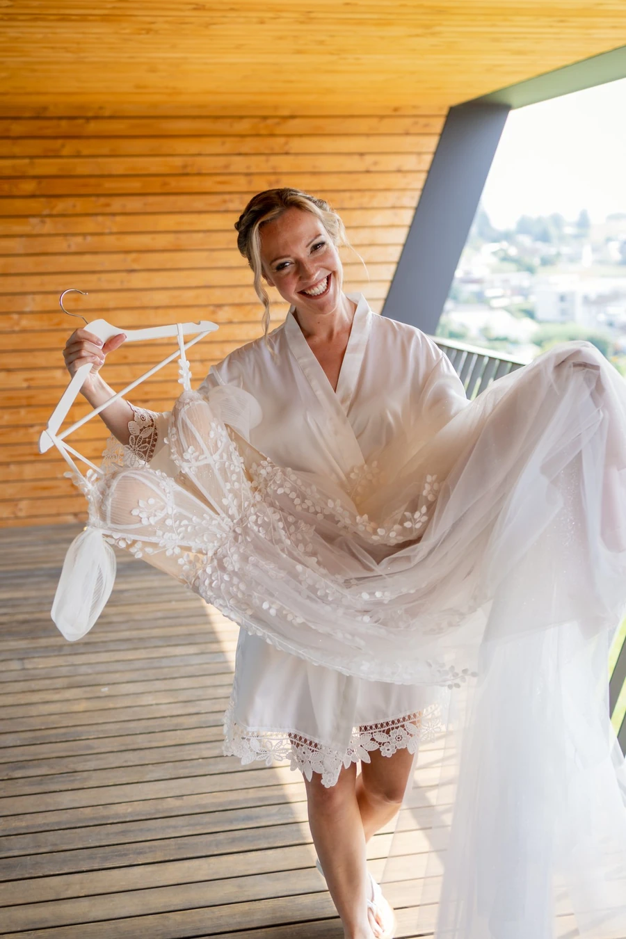 Braut im Morgenmantel hält ihr Kleid am Bügel auf dem Balkon in die Sonne