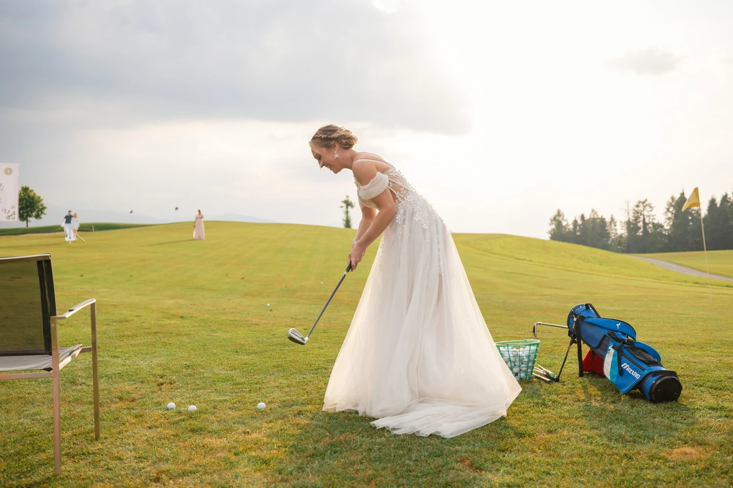 Braut schlägt im Brautkleid auf dem Grün ab, daneben ein Golfbag