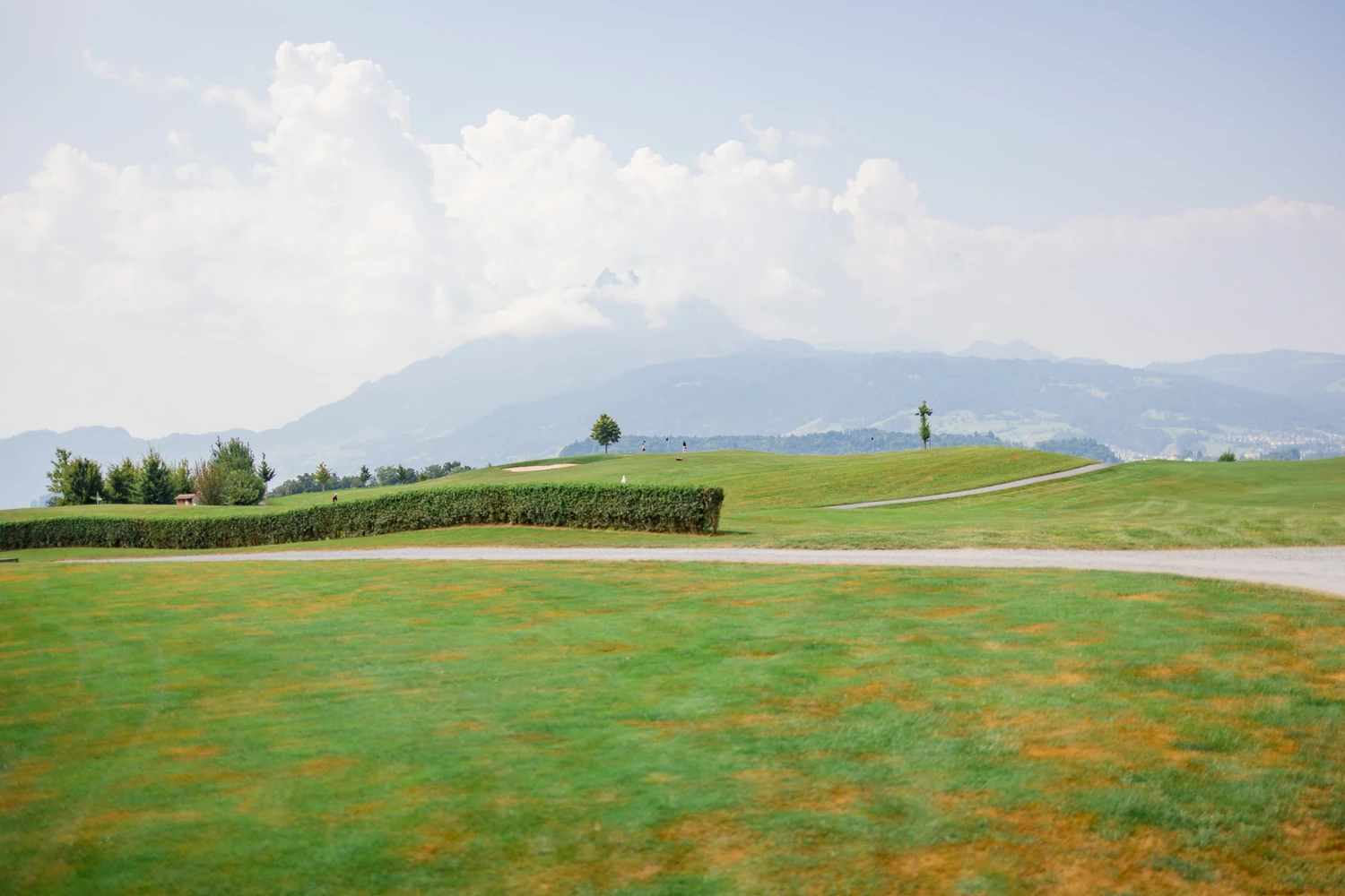 Der Golfplatz mit Hecke, Bunker und Fahne, dahinter Hügel, Berge und eine hohe Wolke
