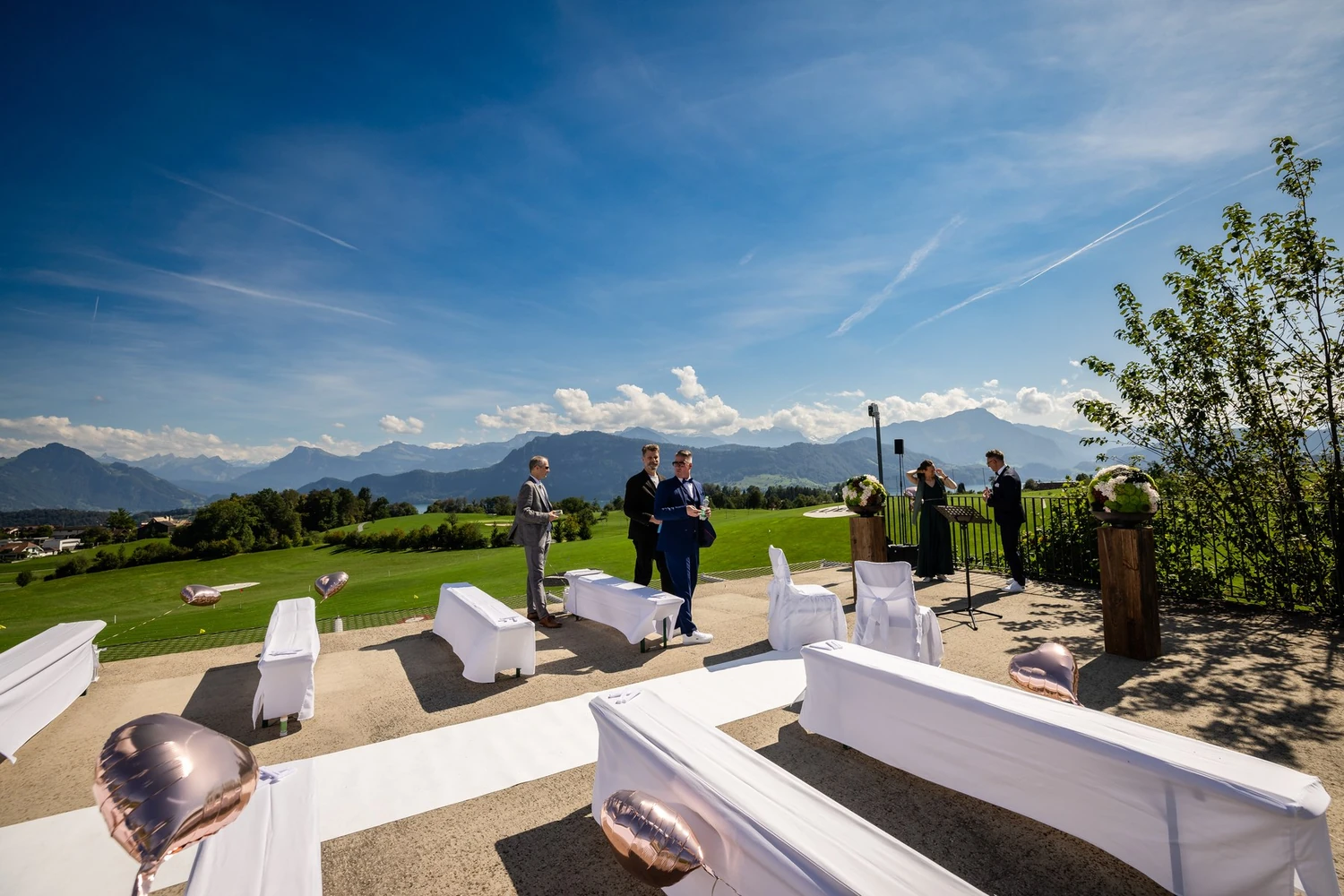 Die aufgestellte Terrasse vor der Trauung mit weissen Bänken, Läufer und Blick über den Golfplatz auf die Berge