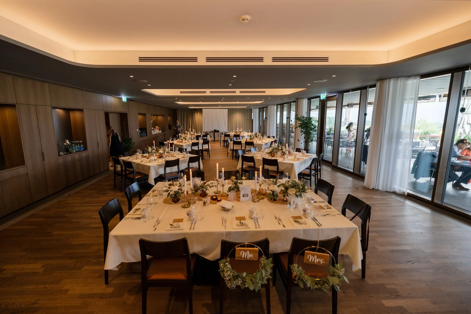 Lange Tafel mit Kerzen und Eukalyptus im Restaurant am Abend, an zwei Stühlen hängen Schilder mit Mr und Mrs