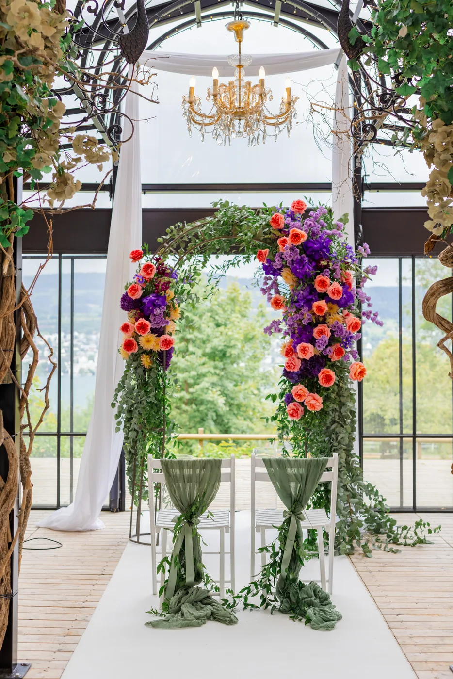 Blumenbogen aus Rosen, Orchideen und Eukalyptus unter dem Glasdach der Pfauenpergola, davor zwei Stühle mit grünen Tüchern