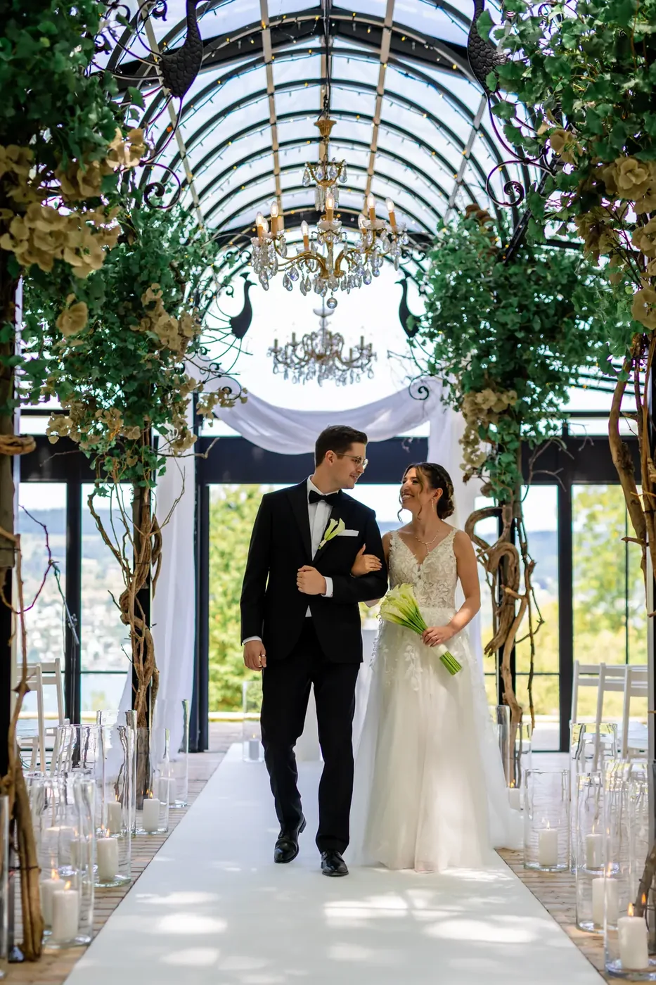 Brautpaar geht Hand in Hand den weissen Mittelgang der Pfauenpergola herunter, dahinter der See