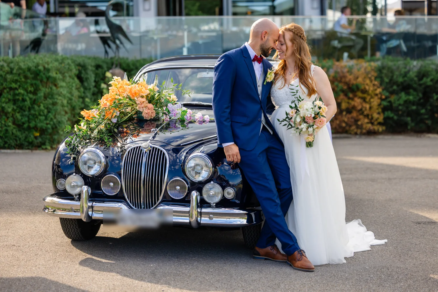 Brautpaar lehnt an einem dunkelblauen Oldtimer, auf der Motorhaube ein grosses Blumengesteck