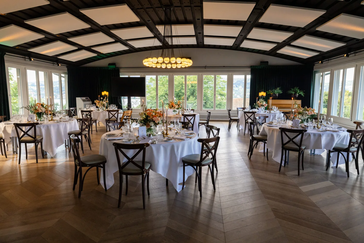 Der AU Saal eingedeckt: runde Tische mit weissen Tüchern, Kerzen und Blumen, ringsum Fenster mit Blick ins Grüne