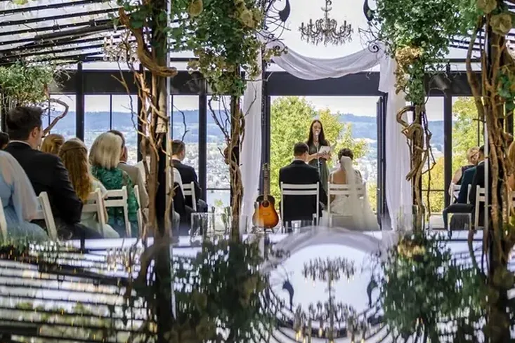 Die Pfauenpergola während der Trauung, gespiegelt in einer Fläche im Vordergrund: Eisenbögen mit Pfauen, Kronleuchter, dahinter der See