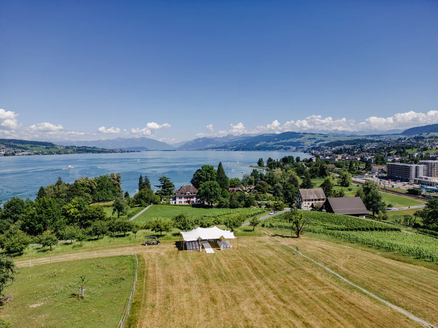 Luftaufnahme: das weisse Stretchzelt steht mit Stuhlreihen auf der gemähten Wiese, dahinter der Landgasthof, der Zürichsee und die Alpen