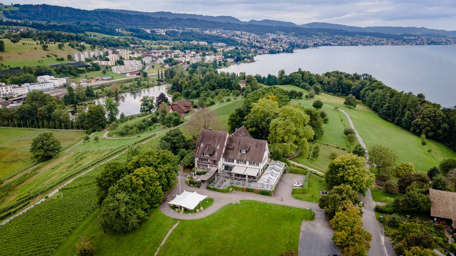 Luftaufnahme des Landgasthofs Halbinsel Au: das weisse Zelt steht auf der Terrasse neben dem Haus, davor die Wiese, dahinter der Zürichsee