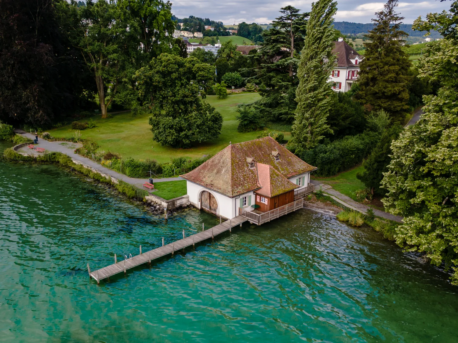 Luftaufnahme eines Bootshauses am Zürichsee mit langem Holzsteg, das Wasser leuchtet türkis