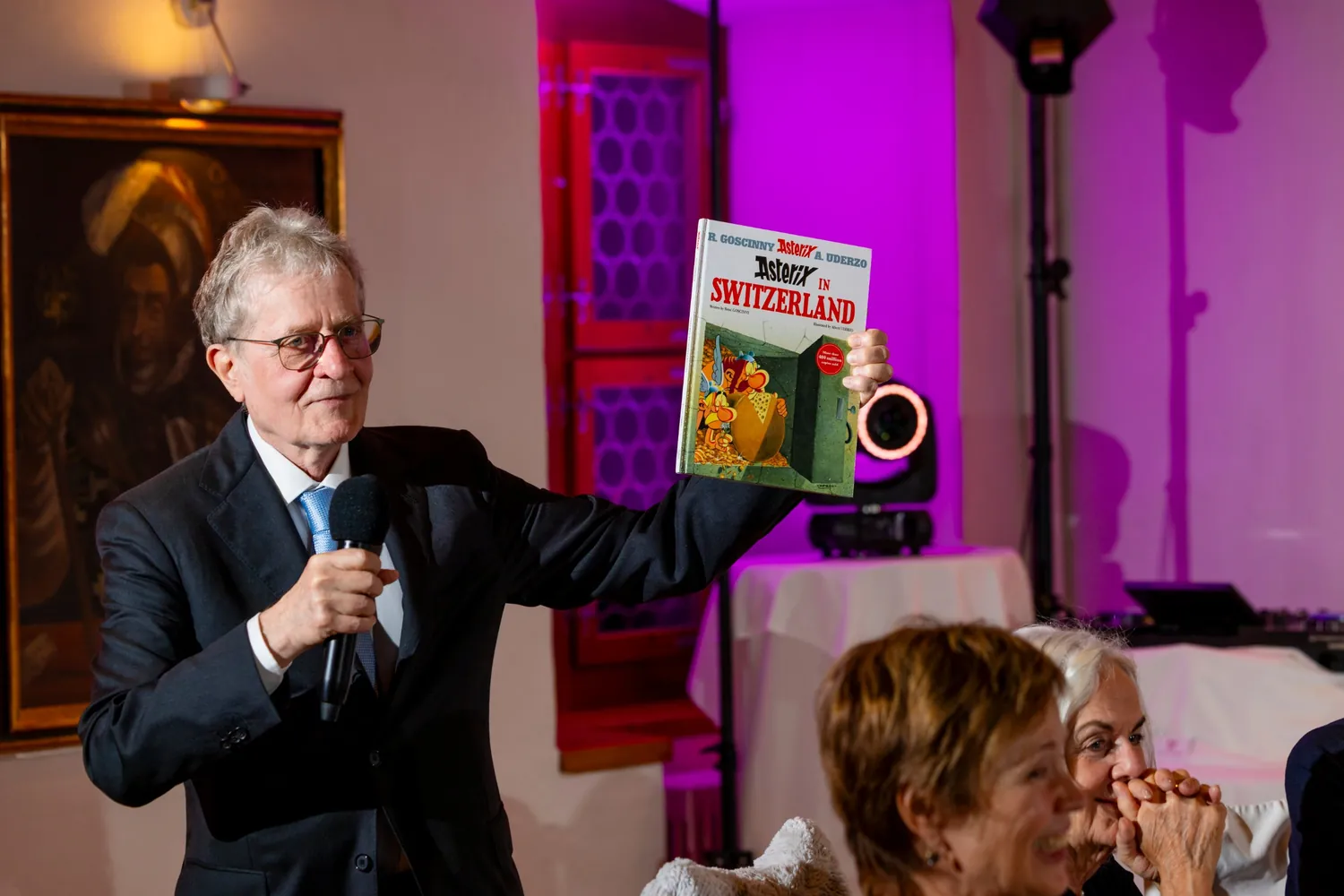 Ein älterer Herr spricht ins Mikrofon und hält bei seiner Rede ein Comicbuch in die Höhe, vorne lachen zwei Frauen
