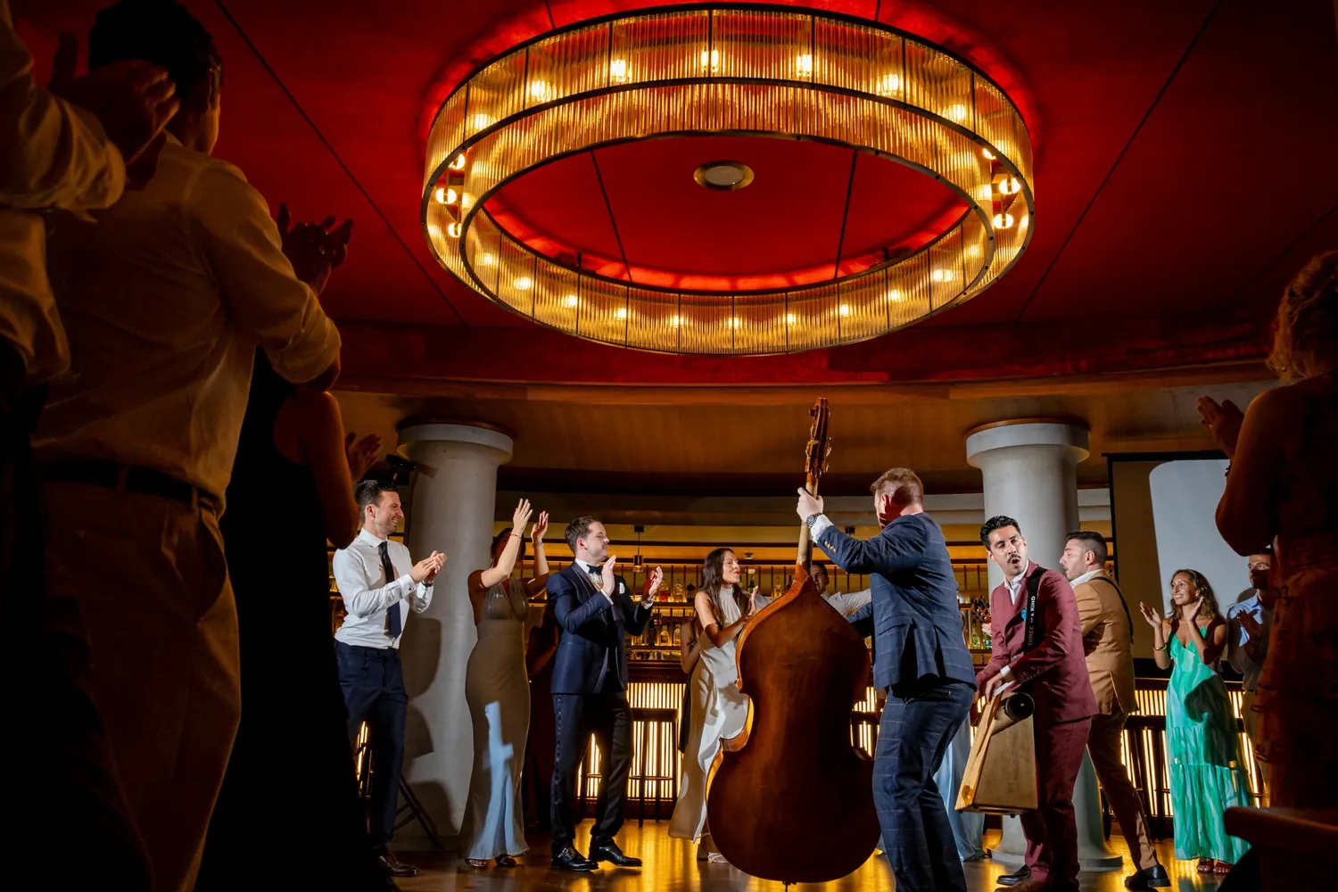 Eine Band mit Kontrabass spielt mitten im Saal unter einem grossen runden Leuchter, die Gäste klatschen im Kreis