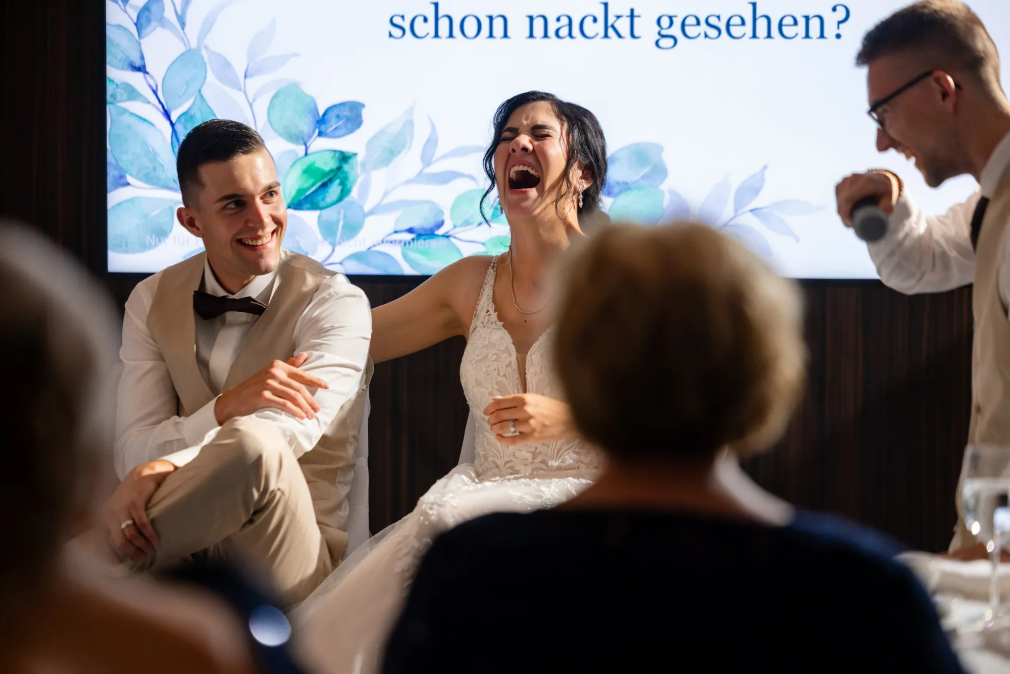 Die Braut lacht laut beim Hochzeitsquiz, der Bräutigam schmunzelt, ein Freund moderiert mit dem Mikrofon, auf der Leinwand steht die Frage