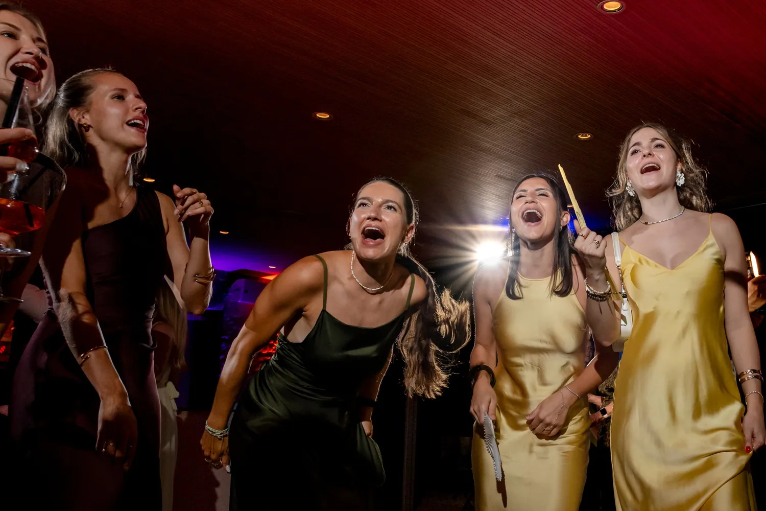 Freundinnen in Seidenkleidern singen auf der Tanzfläche lauthals mit, hinter ihnen strahlt ein Scheinwerfer