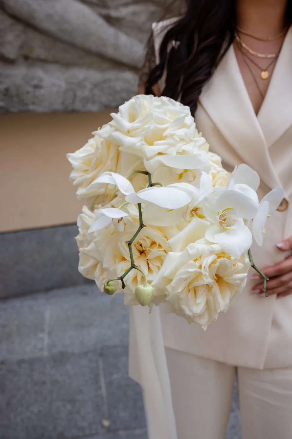 Braut im cremefarbenen Anzug hält einen Strauss aus weissen Rosen und Orchideen
