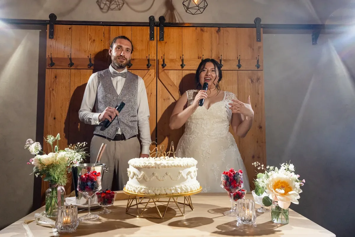 Brautpaar steht mit Mikrofonen am Tortentisch, die Torte noch ohne Beeren