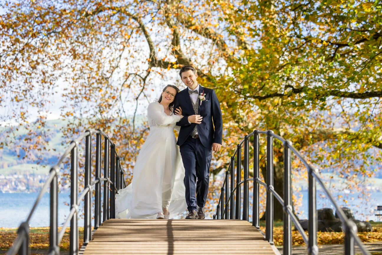 Brautpaar geht über eine Holzbrücke, ringsum herbstlich gefärbtes Laub