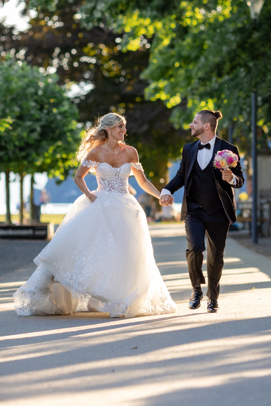 Brautpaar läuft lachend Hand in Hand über eine Promenade, die Braut hält ihr Kleid, Gegenlicht zwischen den Bäumen