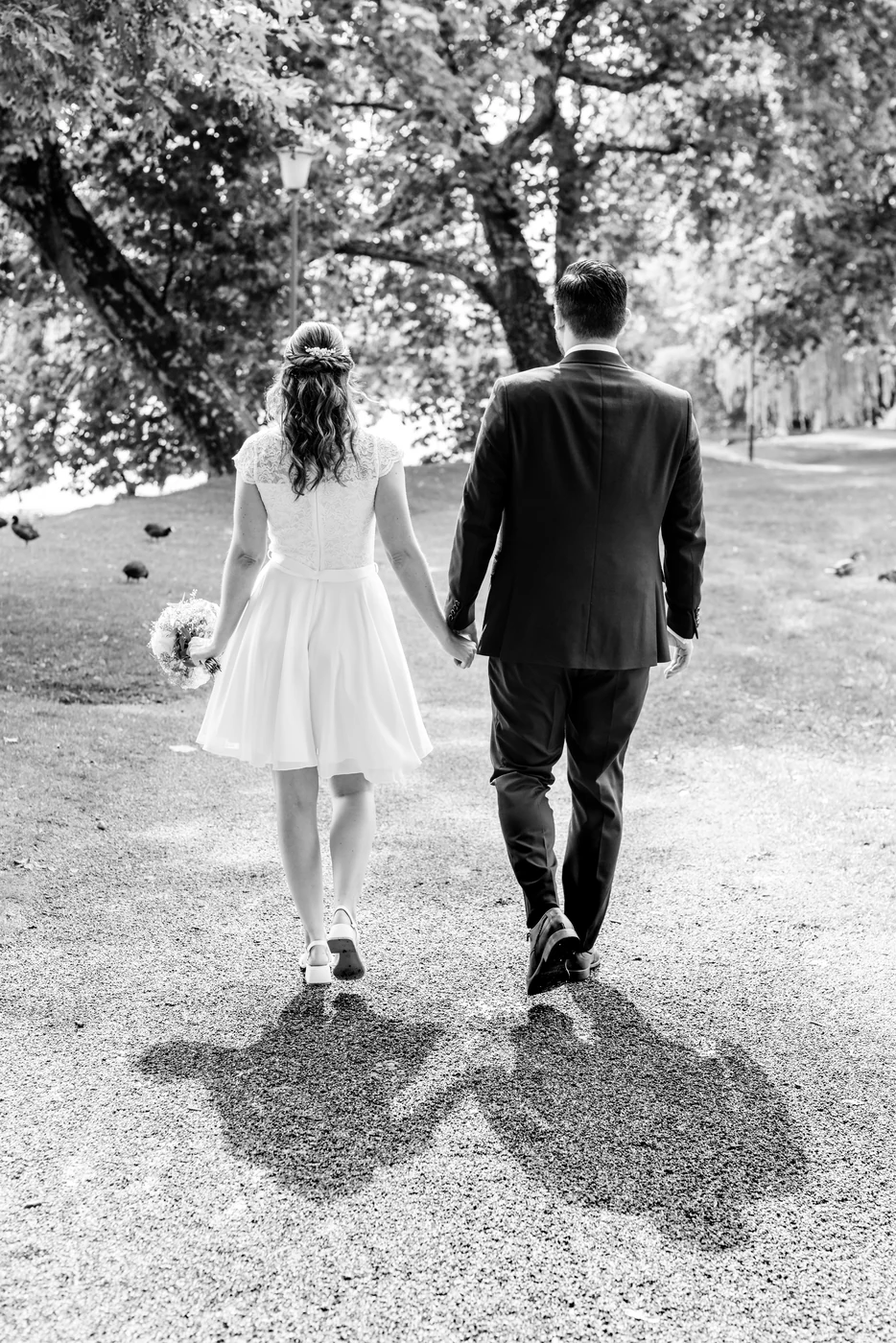 Schwarzweiss: Brautpaar geht Hand in Hand einen Kiesweg entlang, die Schatten fallen lang nach vorne