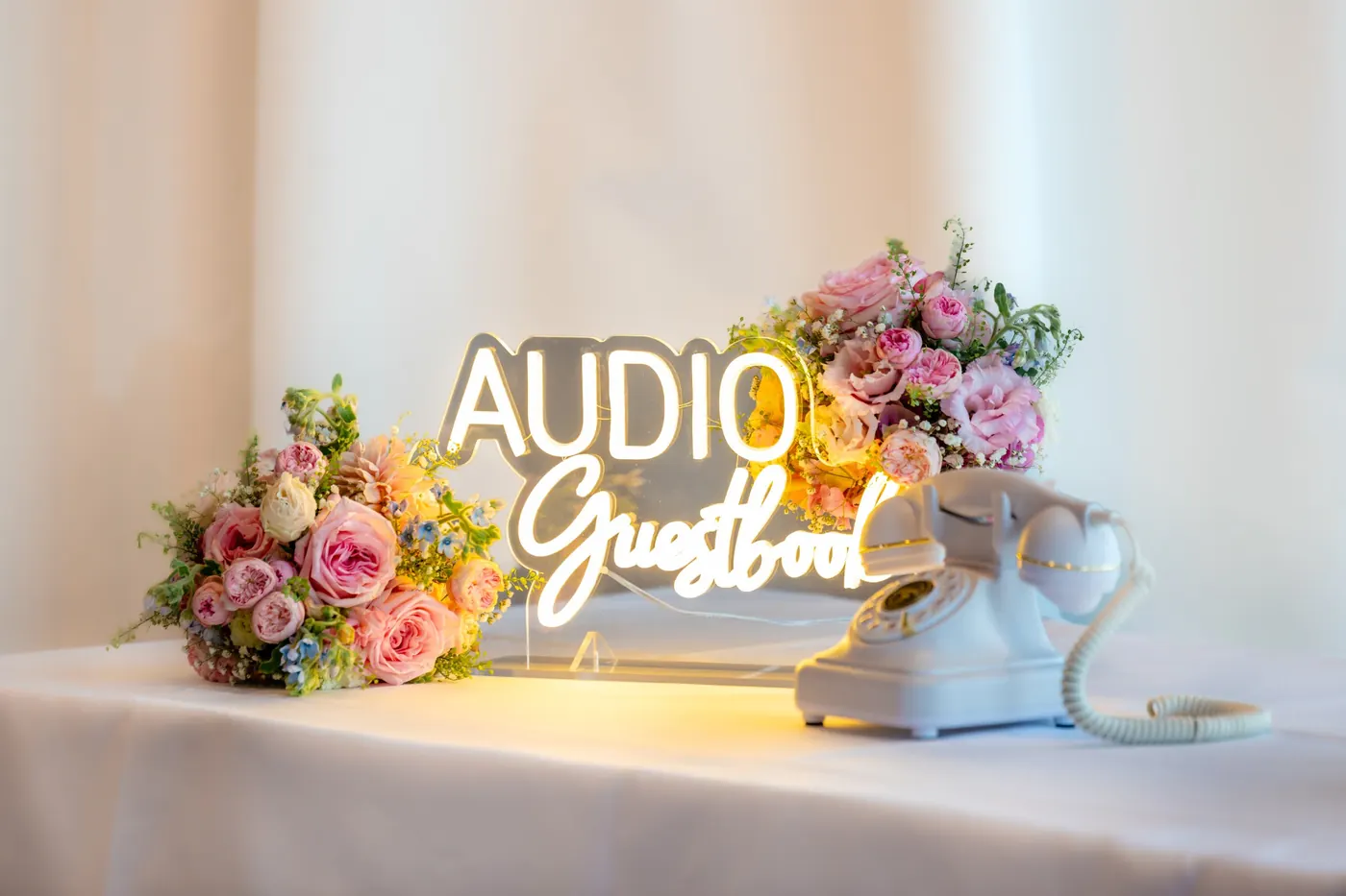 Leuchtschrift «Audio Guestbook» auf einem Tisch, daneben ein weisses Vintage-Telefon und Sträusse aus Rosen