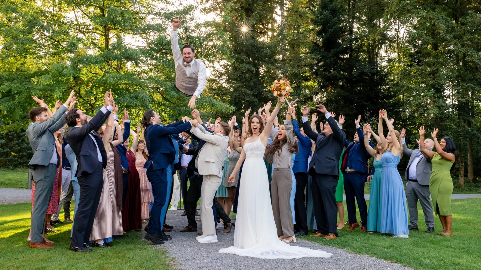Die Gäste werfen den Bräutigam im Park hoch in die Luft, die Braut jubelt mit dem Strauss in der Hand