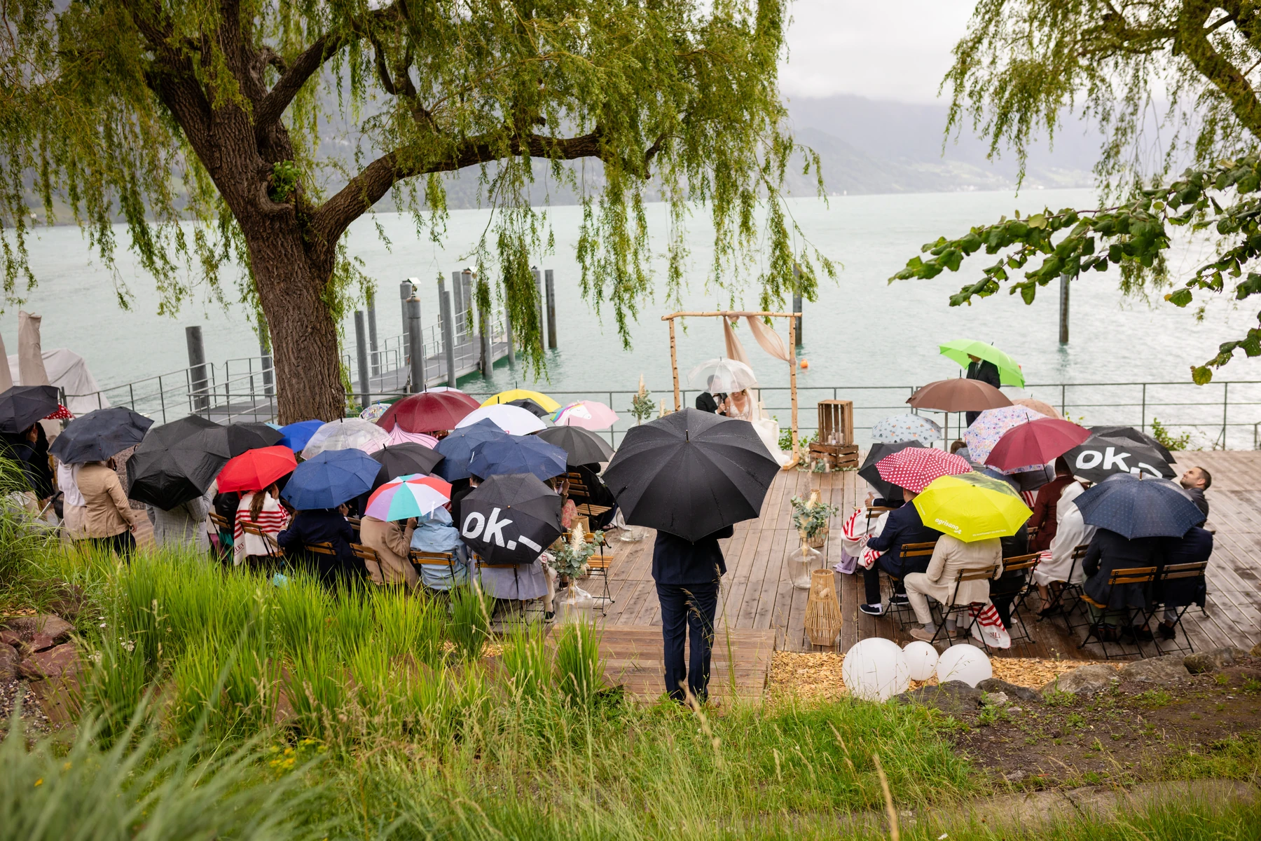 Hochzeitsgesellschaft sitzt bei Regen unter bunten Schirmen auf Holzstühlen an einem See, vorn ein Birkenbogen mit Tuch, im Hintergrund Berge in den Wolken