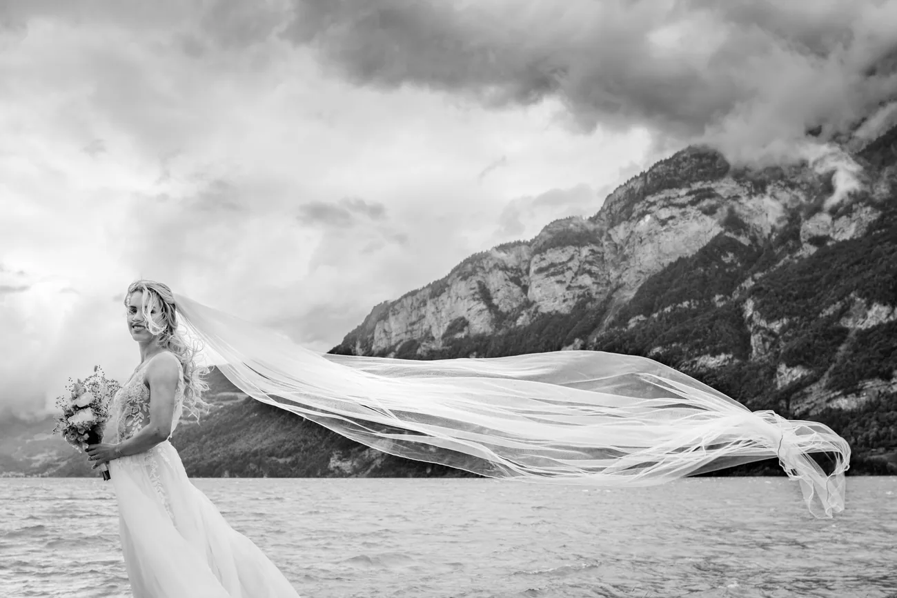 Schwarzweissaufnahme: der Schleier der Braut weht waagrecht über den Walensee, dahinter Wolken über den Bergen