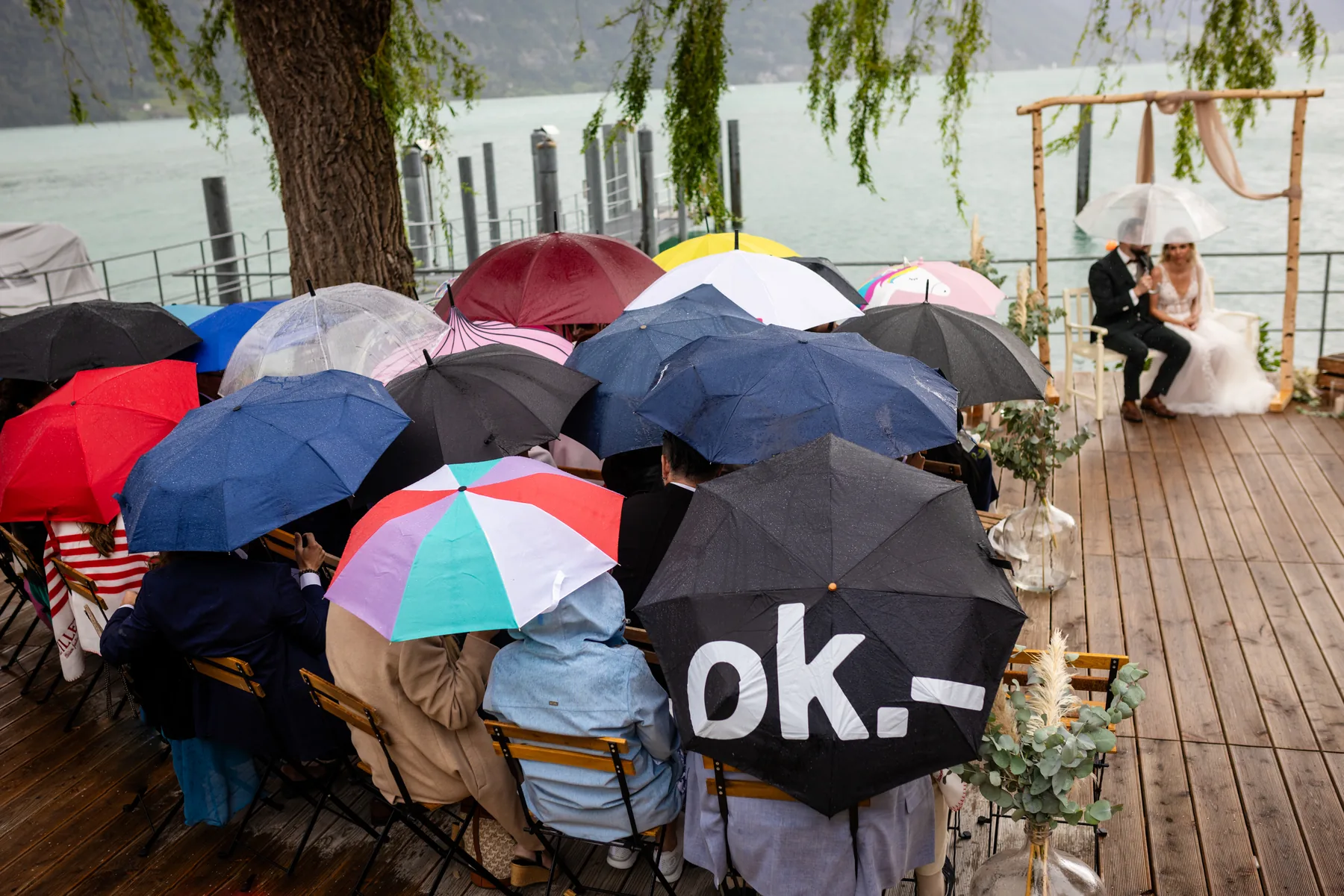 Hochzeitsgäste sitzen unter bunten Regenschirmen, vorne sitzt das Brautpaar unter einem durchsichtigen Schirm vor dem Birkenbogen am Walensee