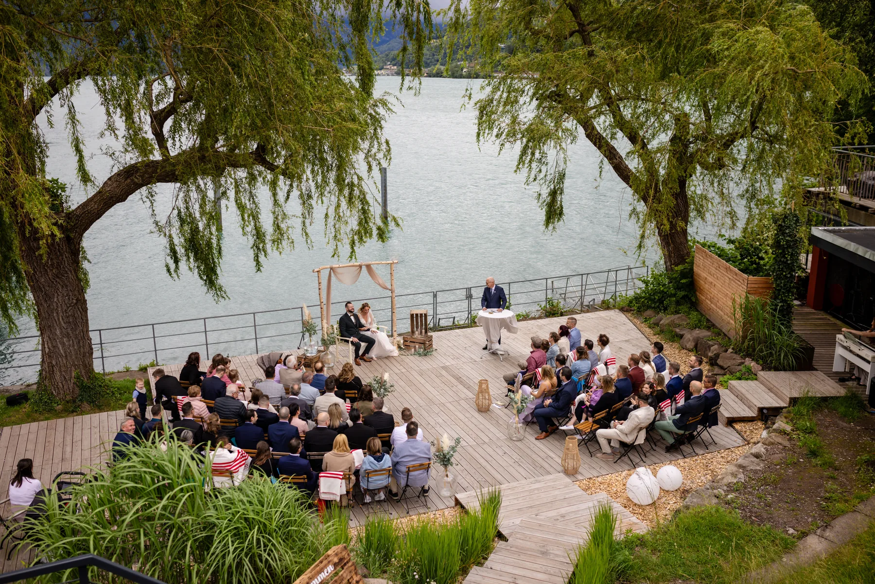 Blick von oben auf die Trauung am Walensee: Gäste sitzen in Stuhlreihen auf einem Holzdeck unter zwei Trauerweiden