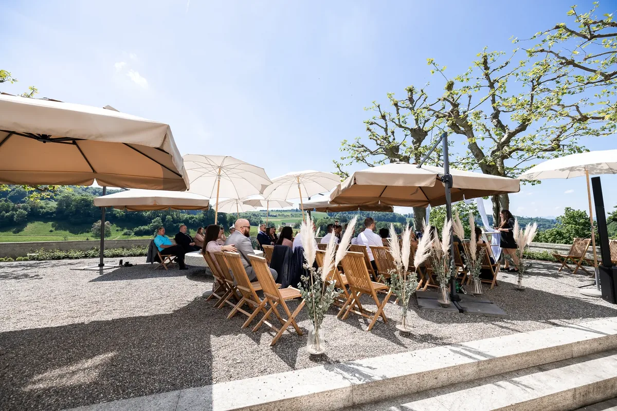Der Zeremonieplatz auf der Terrasse mit Holzstühlen, weissen Sonnenschirmen und Pampasgras, dahinter die Hügel