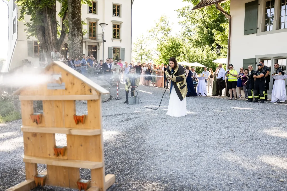 Die Braut in Feuerwehrjacke spritzt mit einem Schlauch auf ein hölzernes Ziel, daneben eine alte Handdruckspritze
