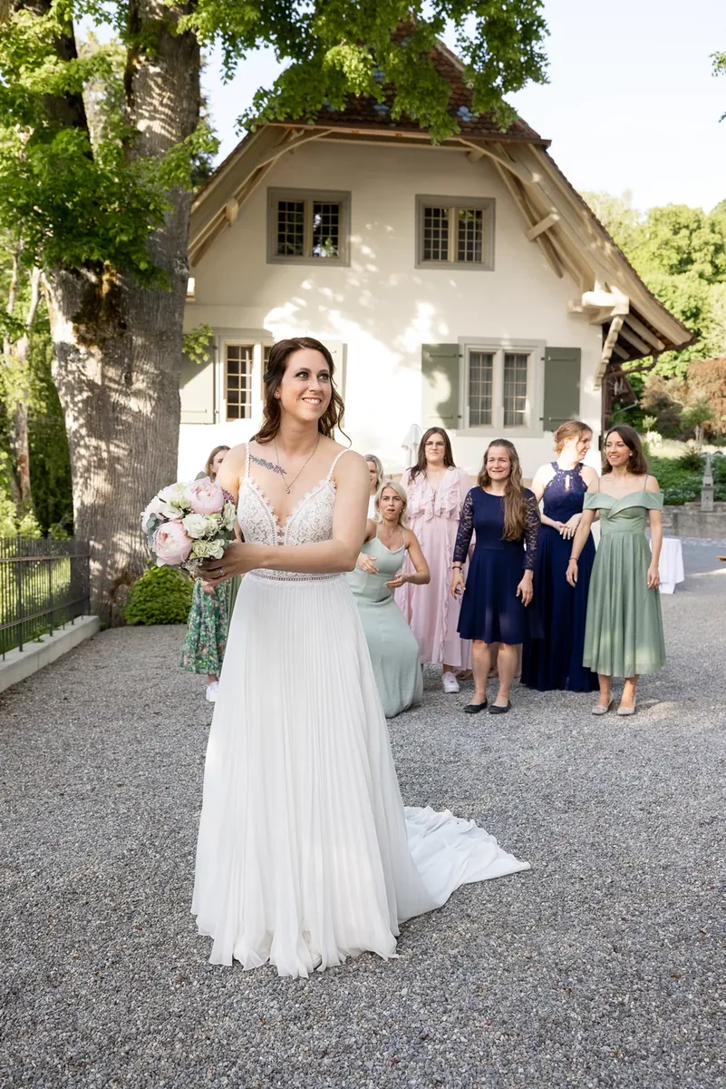 Die Braut mit dem Strauss vor einem hellen Haus mit grünen Fensterläden, dahinter die wartenden Gästinnen