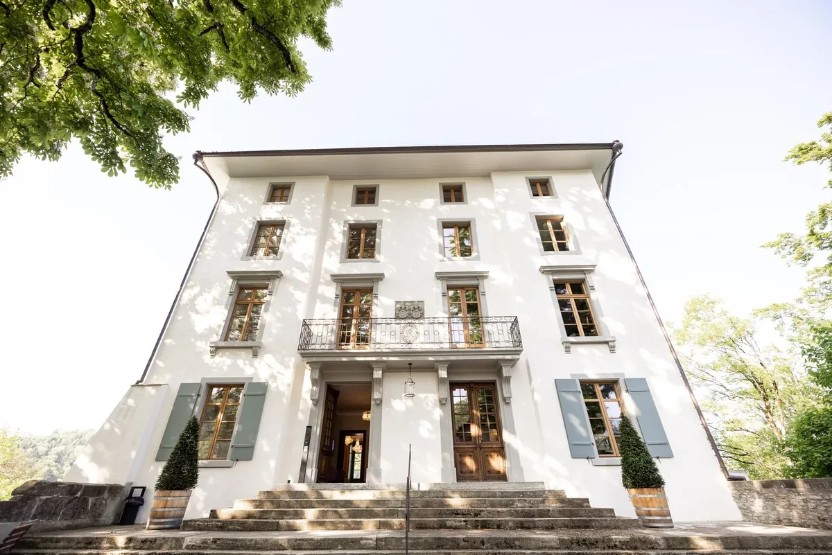 Die helle Fassade des Schlosses Rued mit Freitreppe und alten Bäumen