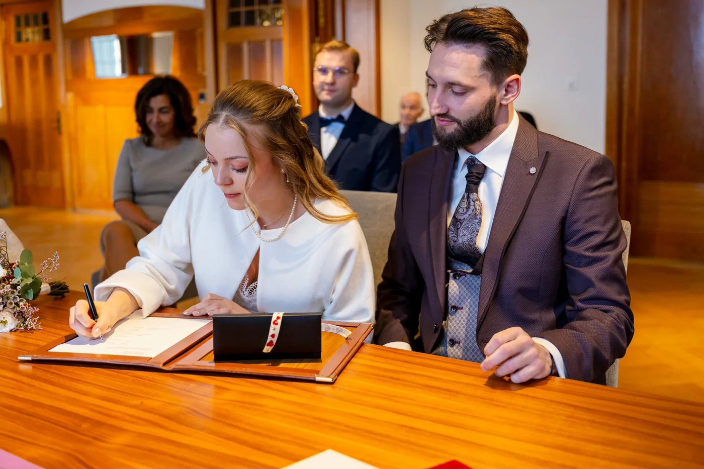 Braut unterschreibt am Tisch im Eheregister, der Bräutigam sitzt daneben