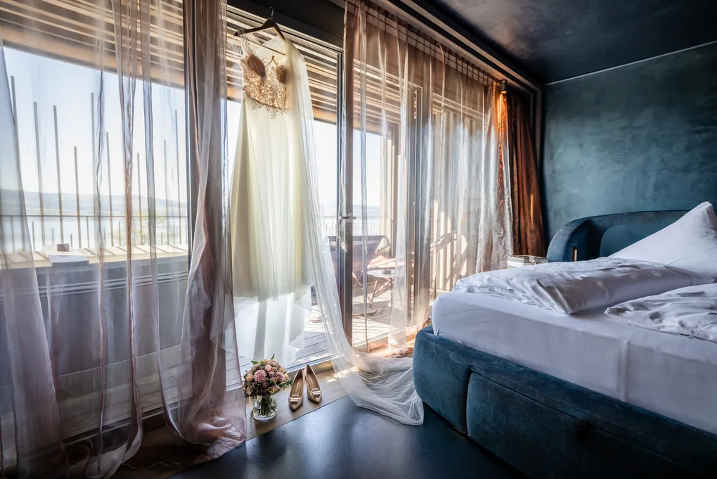 Brautkleid hängt vor dem Fenster eines Hotelzimmers, dahinter der See