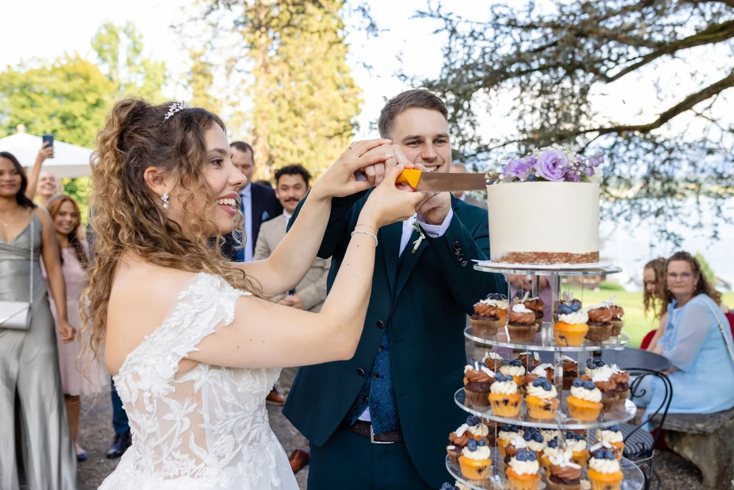 Brautpaar schneidet die Hochzeitstorte im Park an, ringsum die Gäste