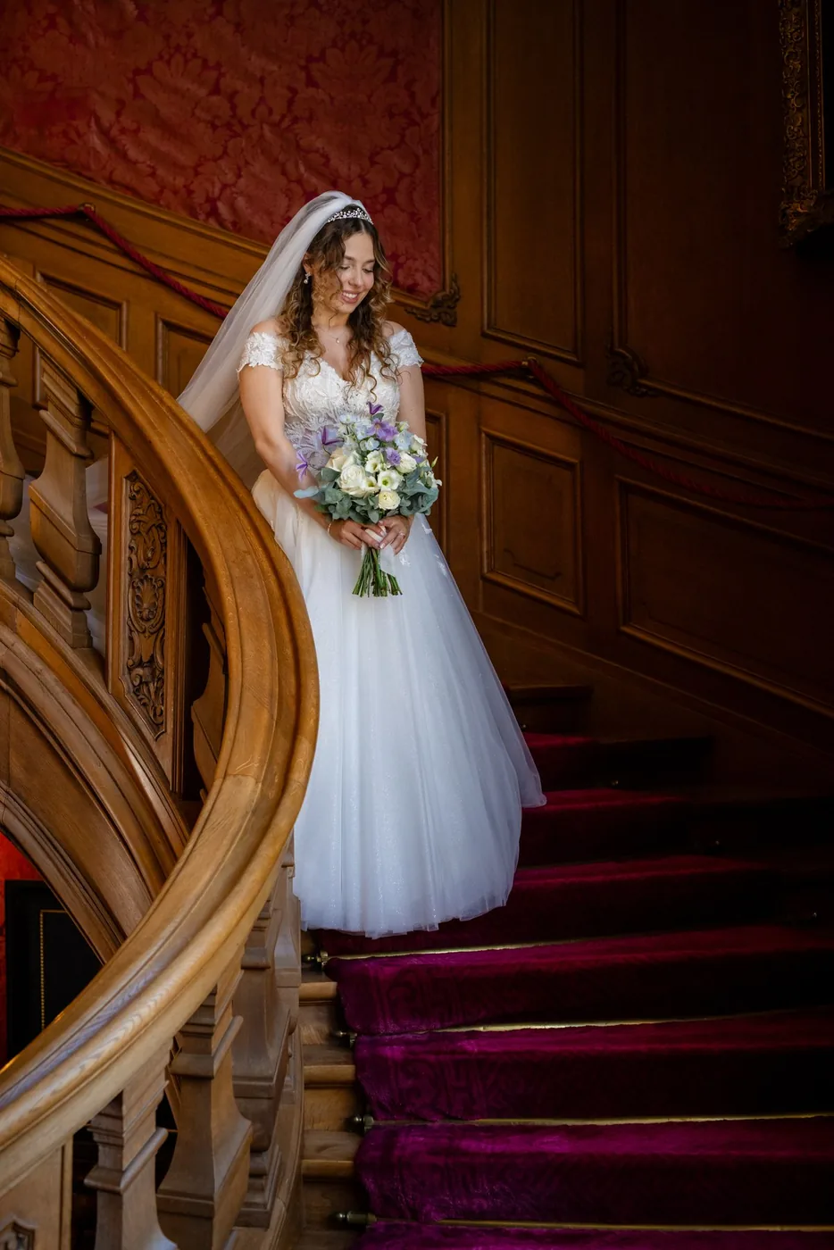 Braut mit Brautstrauss auf der geschnitzten Holztreppe der St. Charles Hall, roter Läufer
