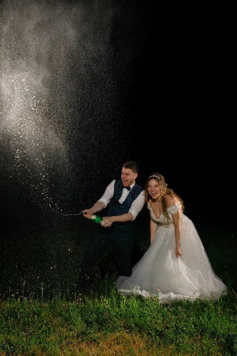 Bräutigam spritzt am Abend Champagner, die Braut lacht, im Dunkeln angeblitzt